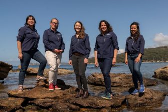 Five women standing on top of a rock in the ocean.