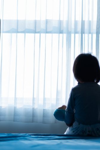 Little baby child sitting on bed playing with the doll in the dark room with light throught curtain
