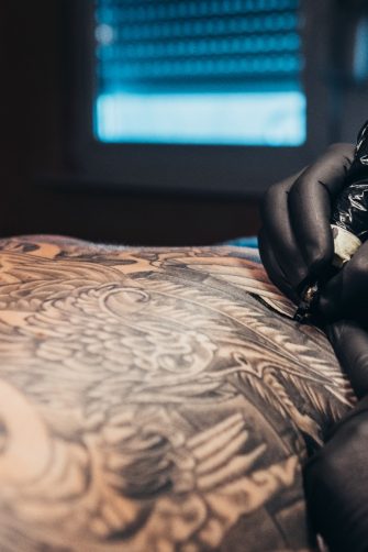 Hands of a tattoo artist wearing black gloves and holding a machine while creating a picture on a man back