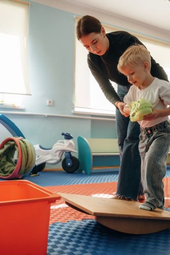 Teacher helping young child balance on a wobble board in a playroom