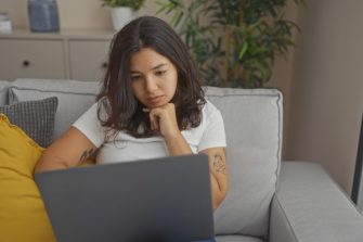 A young hispanic woman in a white shirt using a laptop while sitting on a gray sofa in a cozy living room.