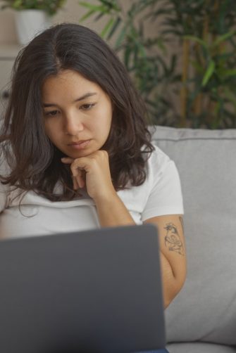 A young hispanic woman in a white shirt using a laptop while sitting on a gray sofa in a cozy living room.