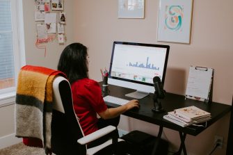 a woman sits at her home workspace, looking at a computer screen 