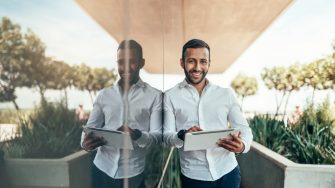 A smiling man with a tablet leans against a reflective wall