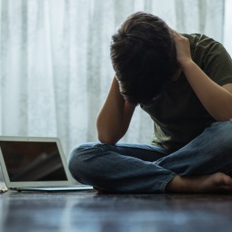 Young person sitting alone next to computer laptop in his room, covering his ears with hands.