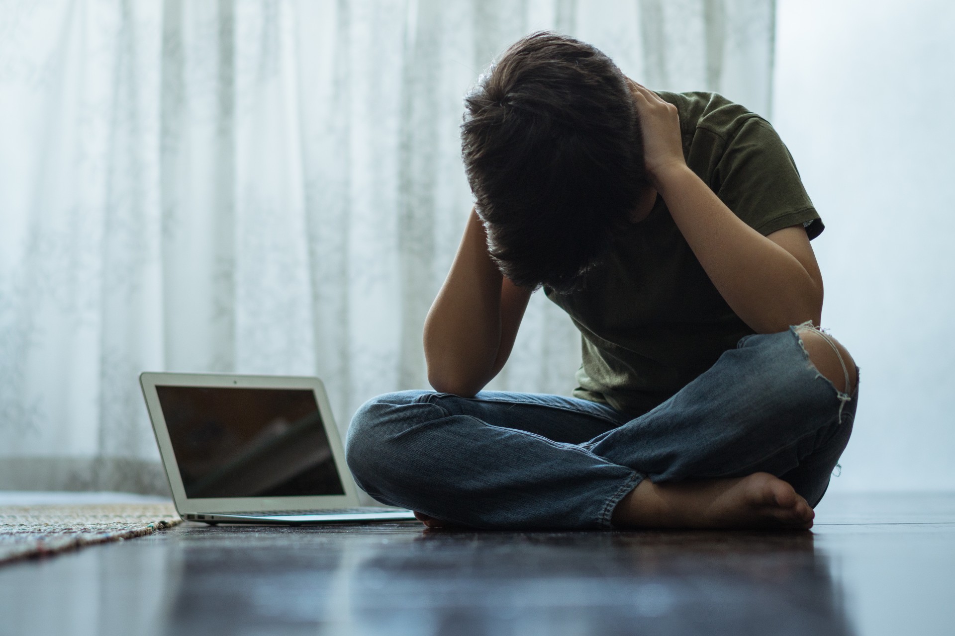 Young person sitting alone next to computer laptop in his room, covering his ears with hands.