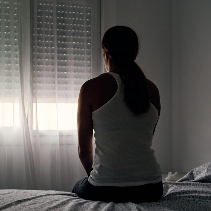 Rear view of an unrecognizable abused woman sitting on her bed looking out the window. Concept of gender violence, domestic violence and depression.