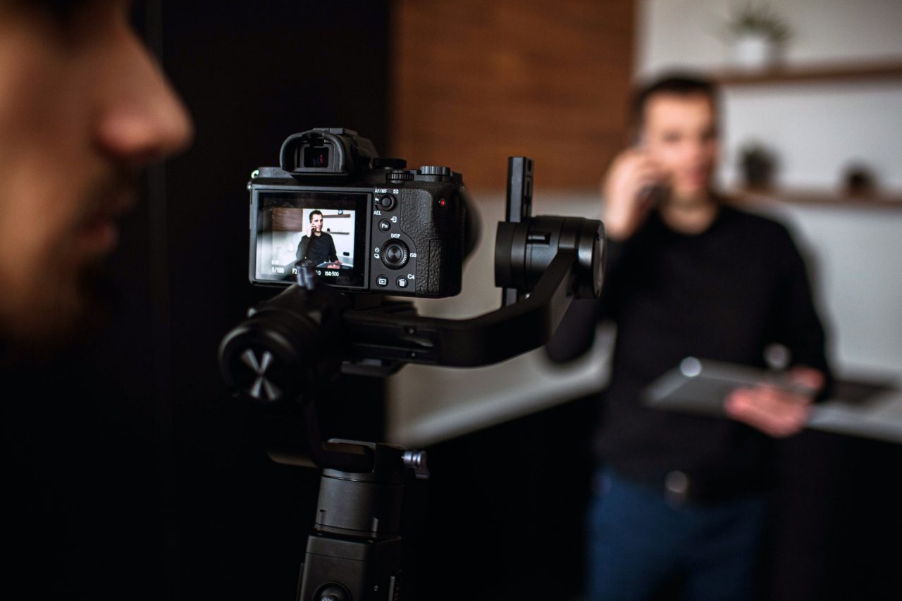 Picture with blurred background where young businessman stand in kitchen and talk on phone. Recording video of dusiness work call or meeting. Camera man stand behind camera and look to right